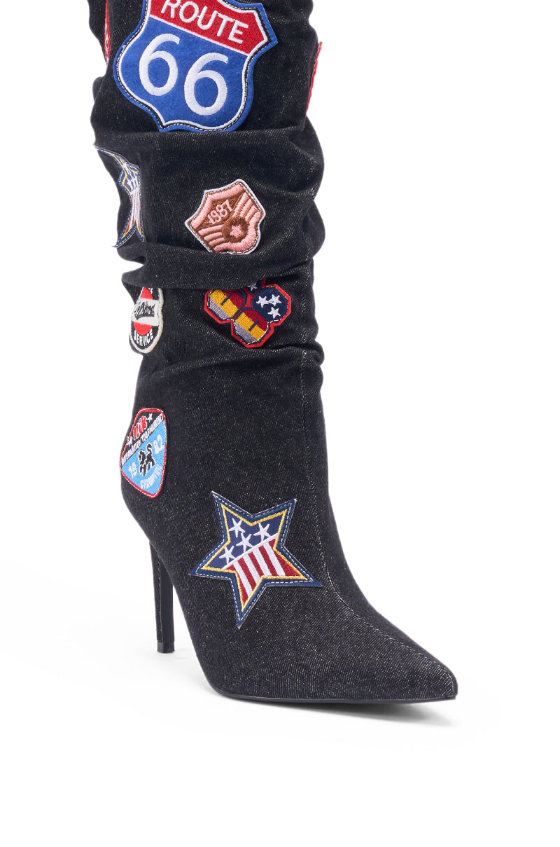 The AZALEA WANG SYLRILAAA DENIM MULTI BOOT is a black denim knee-high boot with a stiletto heel, decorated with vibrant embroidered patches—Route 66, stars, flags, and badges—making it stand out against a white background.