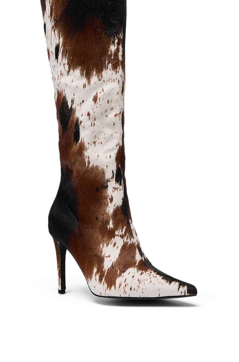 The AZALEA WANG SYREALI COW STILETTO BOOT is a knee-high, pointed-toe boot with a black, brown, and white cow print, featuring a slim stiletto heel and tall faux cow hair shaft. 7