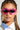 A person with braided hair is wearing distinctive pink CALL ME LATER SUNGLASSES, which boast oval-shaped tinted lenses and a wrap-around plastic frame. They are also accessorized with hoop earrings and a white shirt featuring a blue striped collar, all set against a plain light gray background.