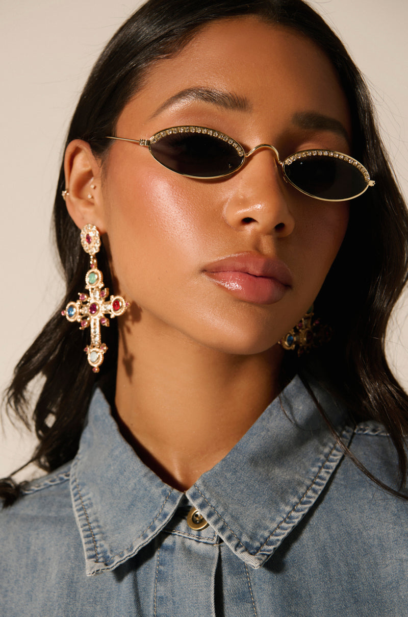 Dressed in a buttoned denim shirt, with long dark hair flowing over their shoulders, a person wears SEE MY DRIP OVAL RHINESTONE SUNGLASSES with gold frames and adjustable nose pads, and ornate cross-shaped earrings adorned with colorful gemstones.