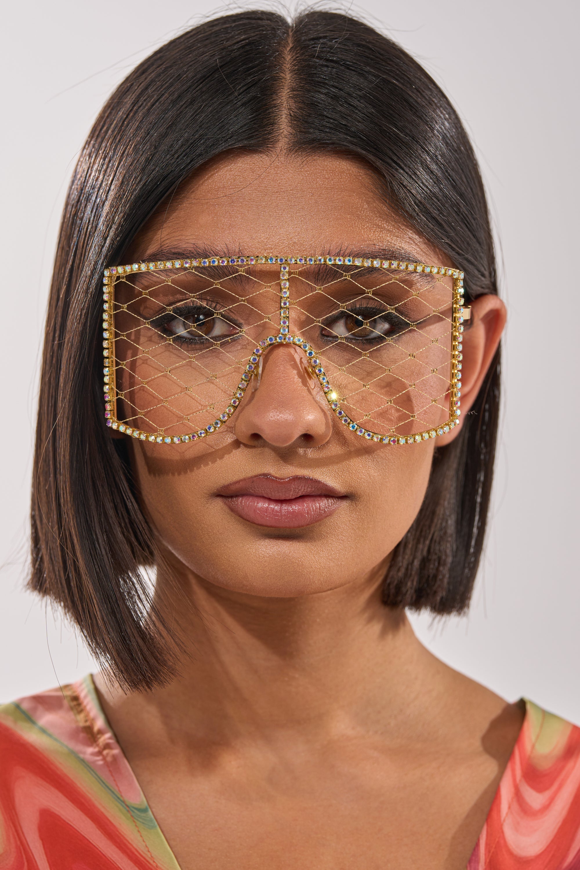 A woman with a sleek bob and neutral makeup wears POP THE BUBBLY GLASSES IN GOLD—rhinestone-embellished metallic frames—paired with a colorful patterned top against a plain light background. 4