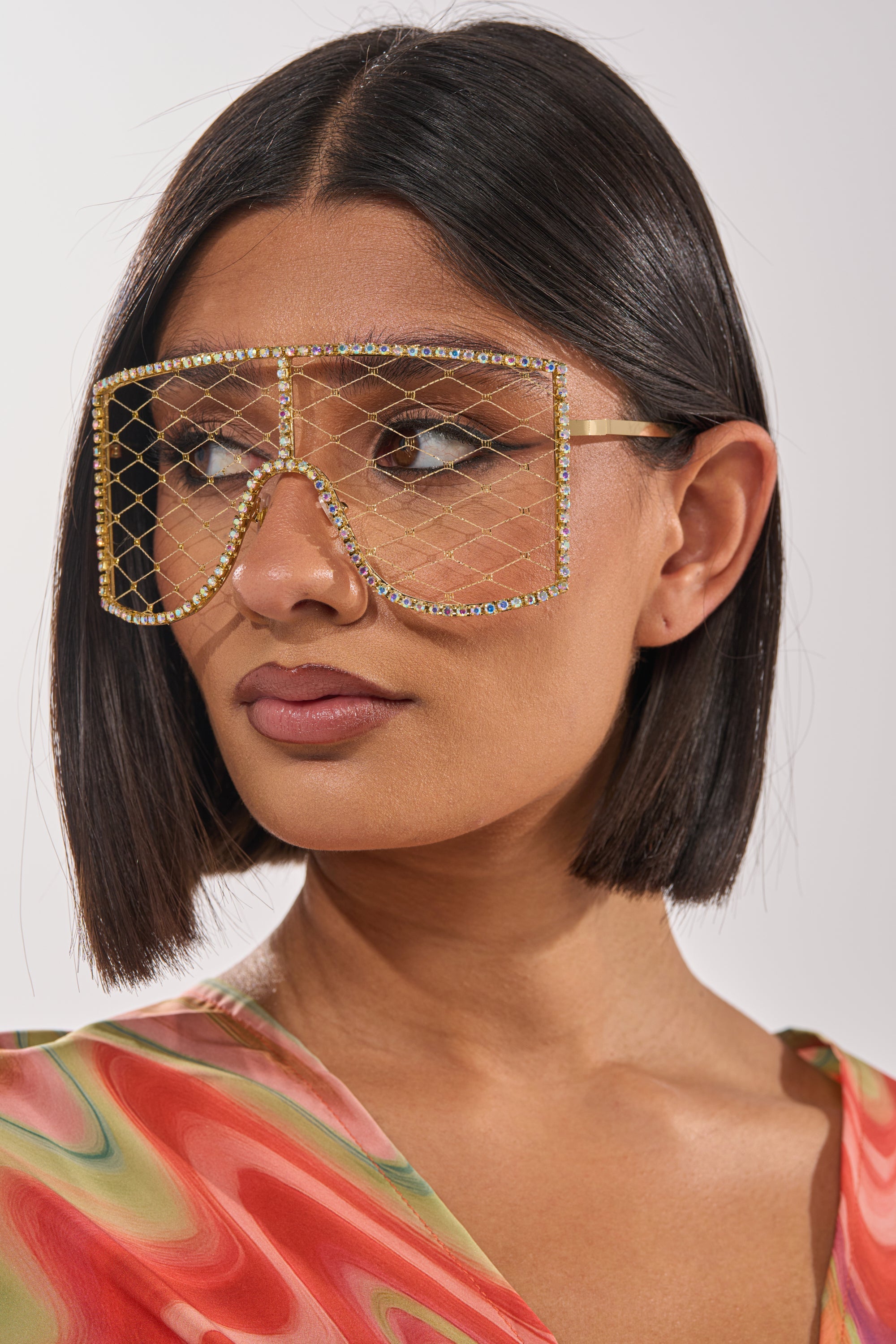 A woman with straight, chin-length dark hair wears POP THE BUBBLY GLASSES IN GOLD—oversized gold-rimmed frames with rhinestone accents. She pairs them with a colorful swirled top and gazes slightly to the side. 5