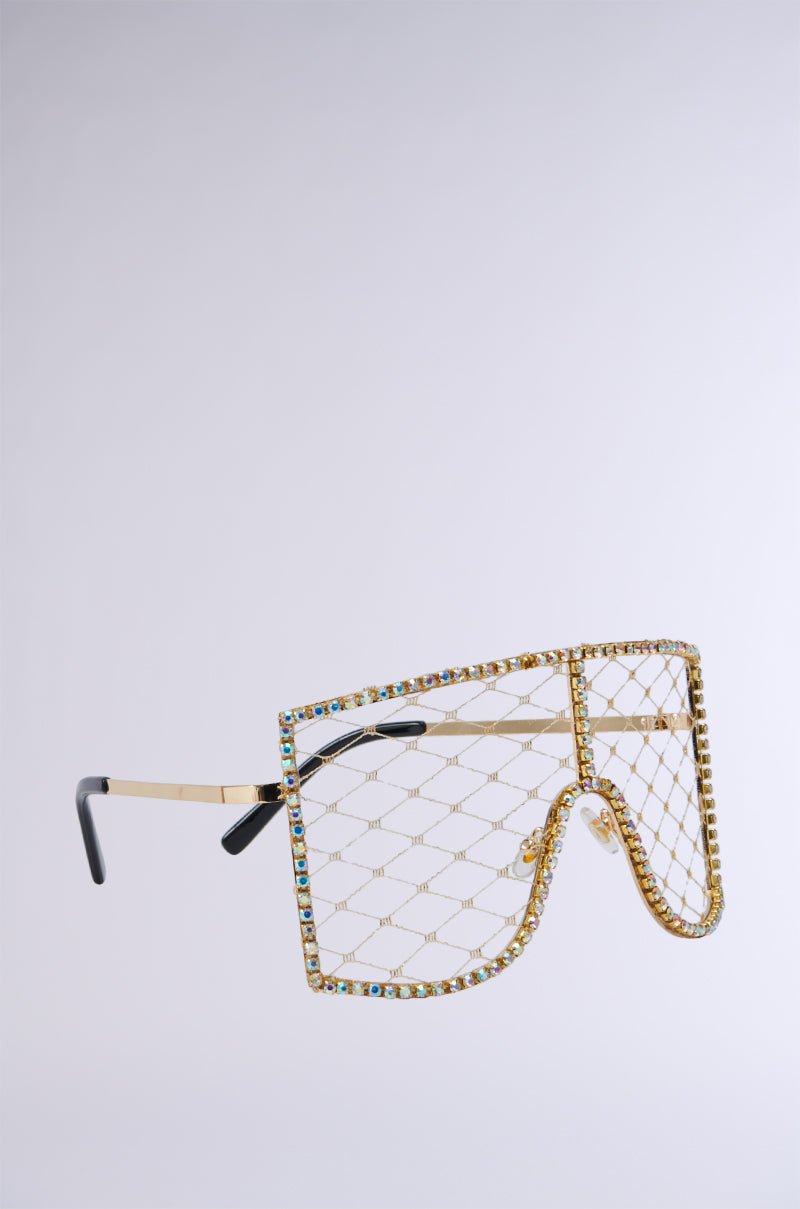 The POP THE BUBBLY GLASSES IN GOLD are luxurious, oversized sunglasses featuring a faux gold metallic frame adorned with rhinestone-embellished detailing and multicolored gemstones outlining the rim. The unique geometric shape paired with black ear tips makes for a bold and extravagant accessory. 6
