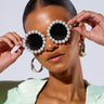 A woman with smooth, pulled-back hair wears oversized RIRI PEARL SUNGLASSES and large hoop earrings. She holds the arms of the stylish lightweight metal frame sunglasses with both hands and is dressed in a mint-green blazer. The background is plain and light-colored.