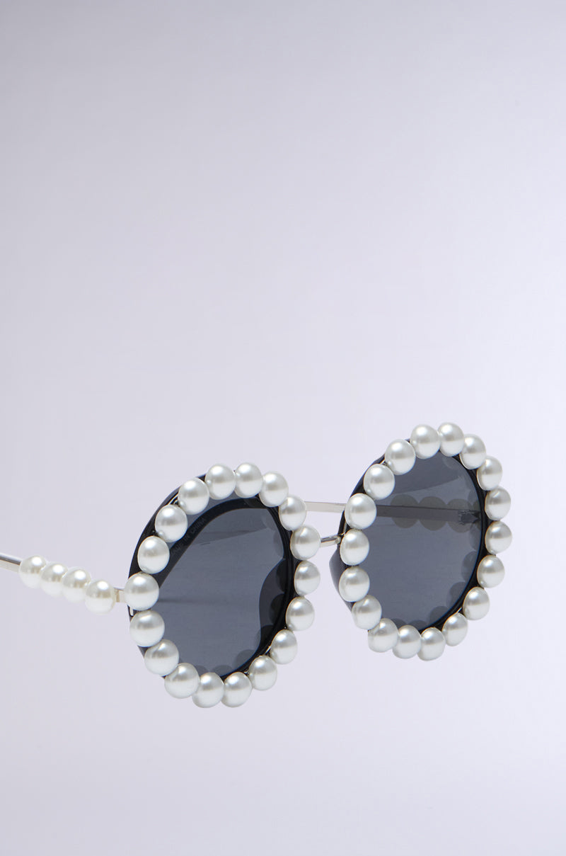 A close-up of the RIRI PEARL SUNGLASSES showcases their round shape and dark lenses. The lightweight metal frame is beautifully adorned with an oversized pearl trim, creating an elegant and stylish appearance. The plain, light-colored background highlights the pearl sunglasses as the focal point.