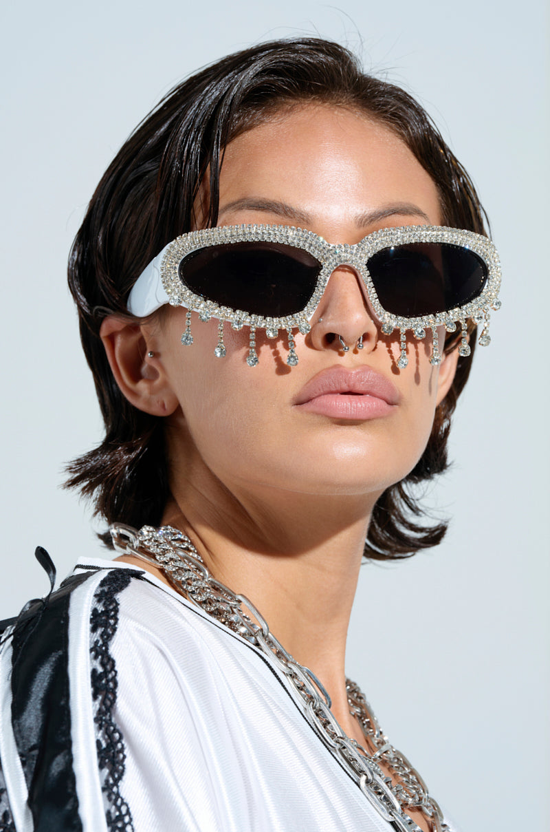 The person flaunts a chic style with short hair and IM YOURS BLING GLASSES, featuring rhinestone trim. A nose ring and thick chain necklace complement the look, while an AKIRA Label white jacket with black accents pops against the light background, showcasing their accessories beautifully.