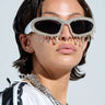 The person flaunts a chic style with short hair and IM YOURS BLING GLASSES, featuring rhinestone trim. A nose ring and thick chain necklace complement the look, while an AKIRA Label white jacket with black accents pops against the light background, showcasing their accessories beautifully.