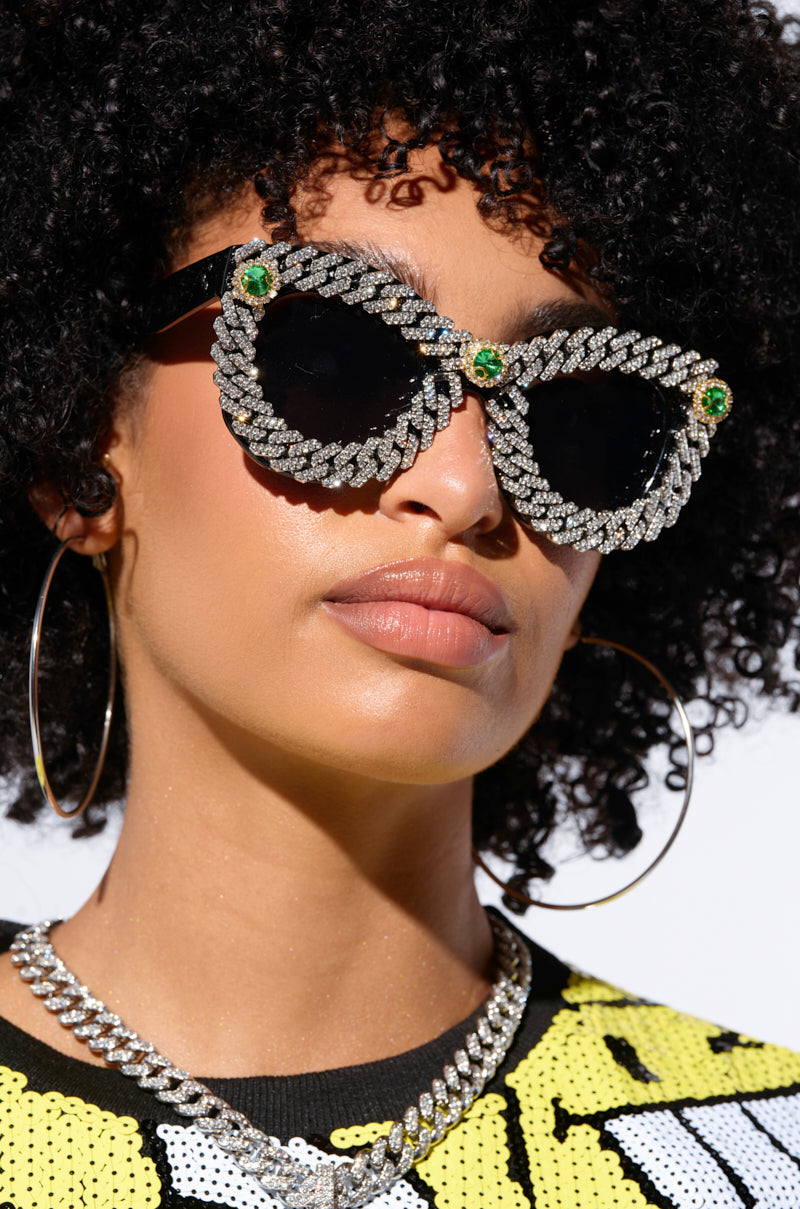 A person with curly hair wears large, black ICE ME OUT SUNGLASSES adorned with intricate, leaf-shaped silver designs featuring green gems at the temples. They also wear a yellow and white top, large hoop earrings, and a silver chain necklace. The background is a plain white.