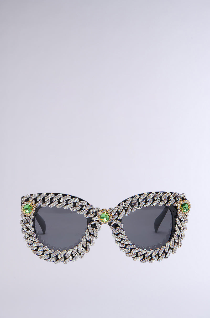 A pair of stylish ICE ME OUT SUNGLASSES featuring dark lenses, an intricately designed frame with a chain-like pattern and rhinestone trim, and prominent green gemstones adorning the corners. The background is plain light gray.