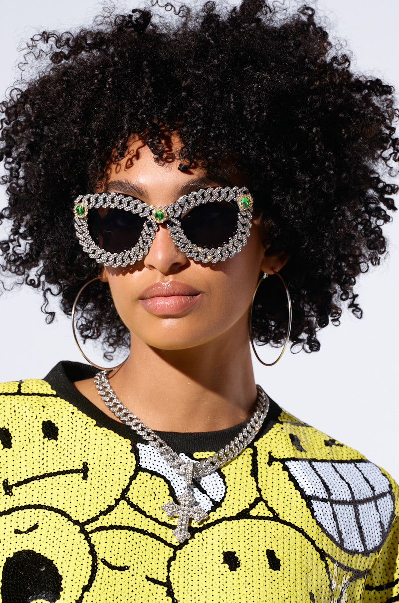 A person with curly hair wears large ICE ME OUT SUNGLASSES featuring gemstone detailing and emerald accents. They have large hoop earrings and sport a yellow top with a pattern of smiling faces. The background is plain white.