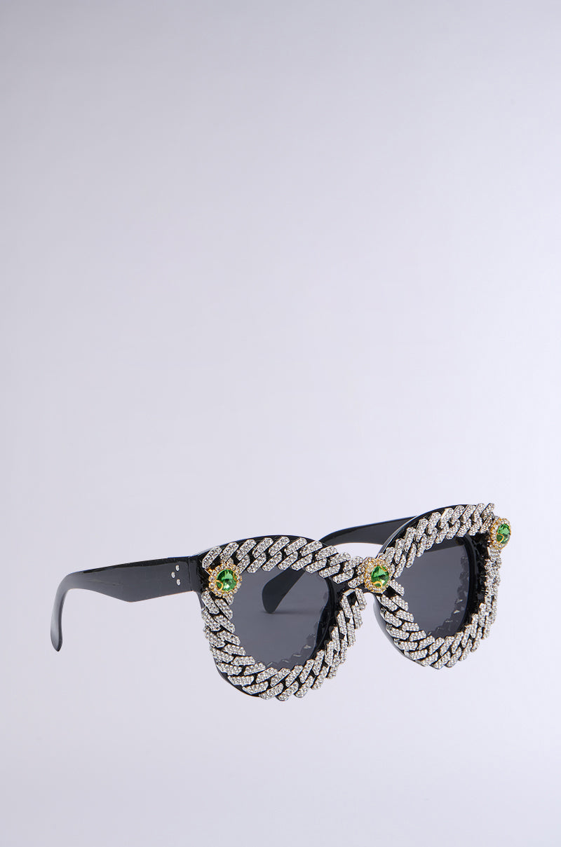 A pair of ICE ME OUT SUNGLASSES with dark lenses, featuring an intricate braided silver frame embellished with a rhinestone trim. Green gemstones at the temples add a vibrant touch and luxurious detail, all set against a light grey background.