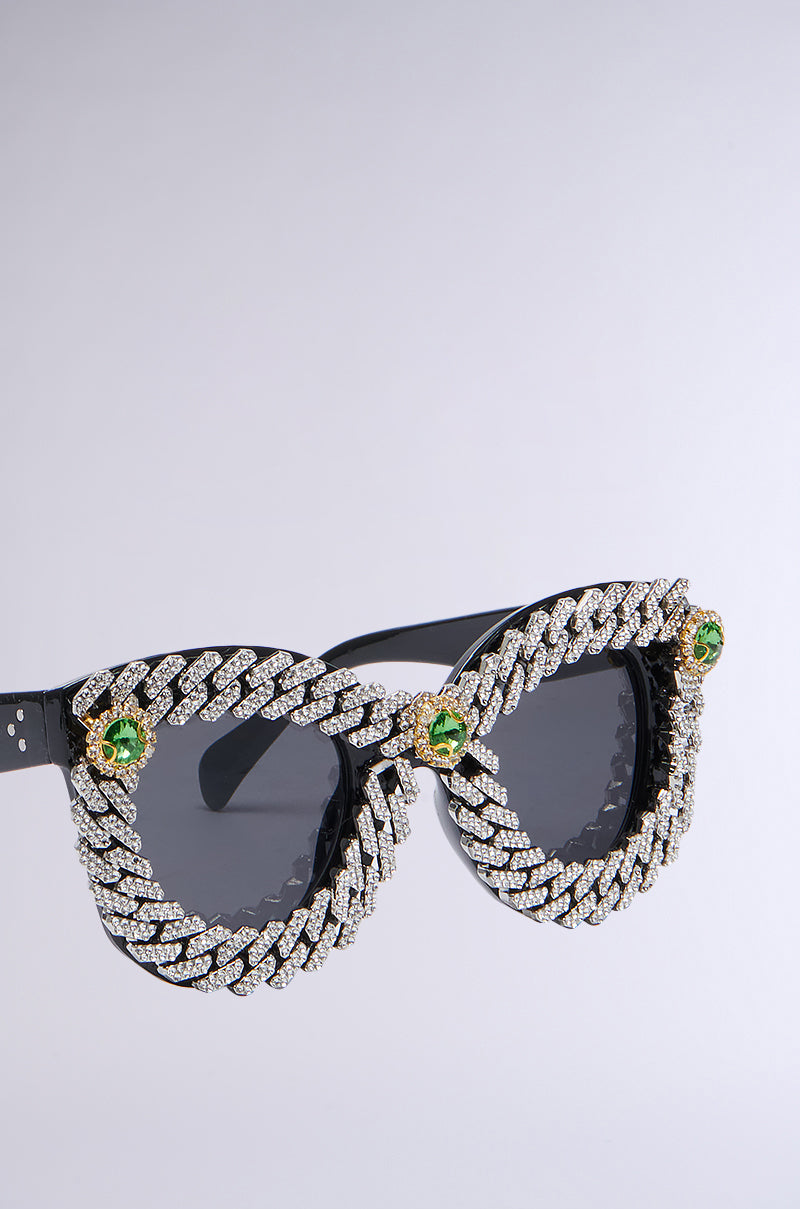 The ICE ME OUT SUNGLASSES feature a pair of black spectacles adorned with a striking, diamond-encrusted chain pattern along the frame. Each corner glistens with green, gem-like stones set in gold, enhancing the luxurious look. The dark-tinted lenses accentuate the sophistication of these sunglasses. They are showcased against a simple, light background.
