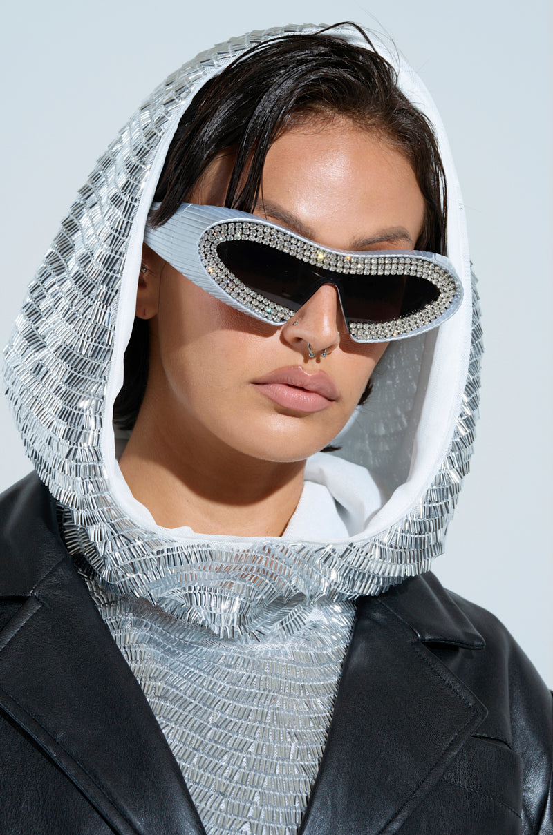 Wearing a shiny, textured silver hood and black leather jacket, a person poses with the TO THE FUTURE BLING GLASSES. The light background highlights their stylish look, creating a unique silhouette.