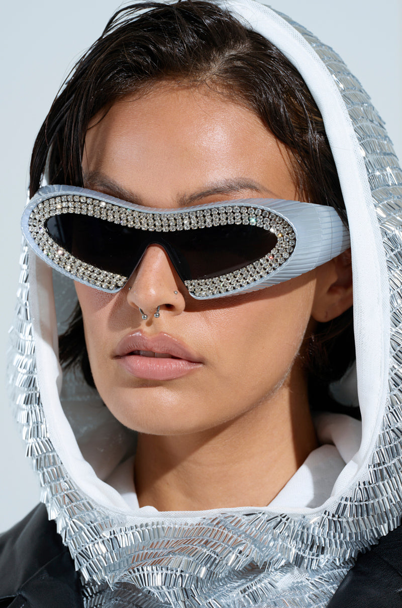 A person with short hair wears TO THE FUTURE BLING GLASSES and a shiny, metallic-textured hooded garment. They have facial piercings and a neutral expression against a plain background.