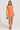 A person confidently stands in the INTO YOU SLINKY MINI DRESS, an orange, sleeveless bodycon with a racerback design. They hold a small, patterned handbag and wear strappy, embellished heels. The neutral background perfectly highlights their stylish ensemble.