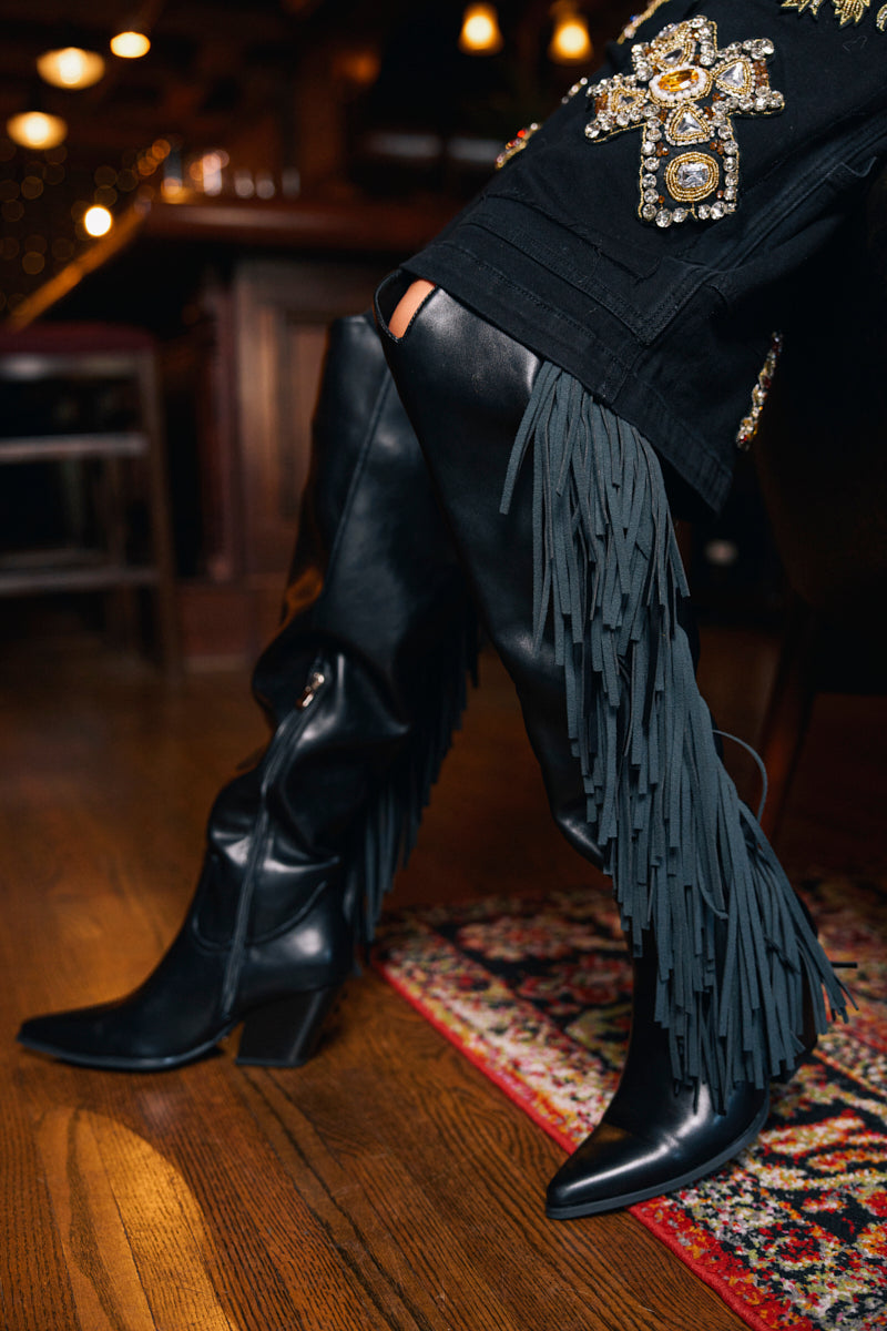 Someone sits indoors on a wooden floor, wearing black pants with embellished patches and the AZALEA WANG SZYMON BLACK WESTERN BOOT WITH FRINGE DETAILING; a patterned rug is partly visible, finishing the stylish look.