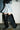 Person wearing a flared, frayed denim skirt, black ribbed sweater, and AZALEA WANG SZYMON Black Western Boot with Fringe Detailing, standing indoors on a concrete floor against a white brick wall.