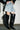 A person stands against a white wall, wearing a flared frayed-edge denim mini skirt and the AZALEA WANG SZYMON BLACK WESTERN BOOT WITH FRINGE DETAILING. Dramatic shadows fall across the textured floor.