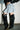 Wearing a flared light-wash denim mini skirt and the AZALEA WANG SZYMON Black Western Boot with fringe detailing, someone stands against a white brick wall and concrete floor.