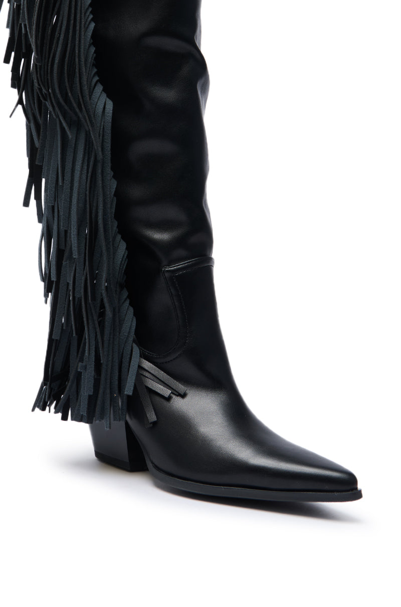 A close-up of the AZALEA WANG SZYMON BLACK WESTERN BOOT showcases its pointed toe, block heel, and long black fringe along the side, combining knee-high style with a Western-inspired cowgirl vibe.