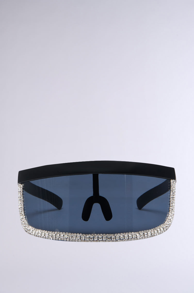 The CLEAR VIEW VISOR SUNGLASSES boast an oversized visor silhouette with a dark rectangular lens framed by a wide, black border. The bottom edge of the frame is elegantly embellished with a single row of sparkling rhinestones, set against a subtle, light gradient background.