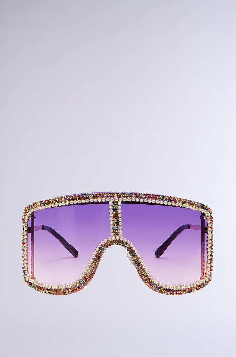 The IN YA CITY BLING SUNGLASSES are a pair of oversized shield sunglasses with a single purple ombré lens. The frame is adorned with colorful rhinestones, forming a sparkling border around the lens. The arms of these bling sunglasses are sleek and black, contrasting against a plain, light backdrop.