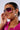 A person wearing IN YA CITY BLING SUNGLASSES with purple ombré lenses and large gold hoop earrings. Their hair is pulled back, and they are dressed in a shiny, bright pink garment. They touch the side of their oversized shield sunglasses with one hand.