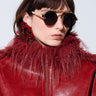 A person wearing CATCH ME STUD SUNGLASSES, featuring round lenses with spike embellishments and a gold metal frame, along with a red leather jacket adorned with a furry collar and red earrings, stands against a light background.