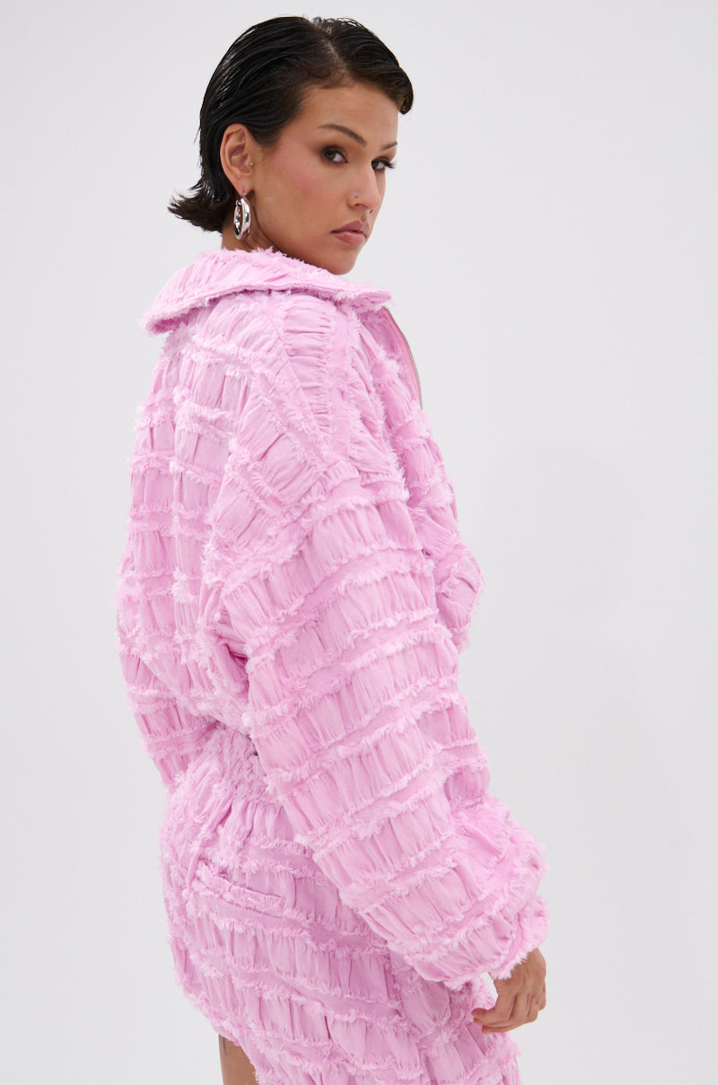 A woman with short dark hair poses in the BUBBLEGUM BUBBLE HEM BOMBER, a textured light pink coat with a belted waist and large collar. She turns slightly to the side, looking over her shoulder against a plain light background.