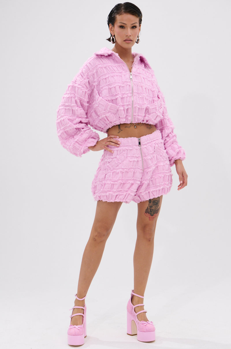 A woman poses confidently in the BUBBLEGUM BUBBLE HEM BOMBER, styled with a matching mini skirt and pink platform heels. Her slicked-back hair and visible thigh tattoo stand out against the plain white background.