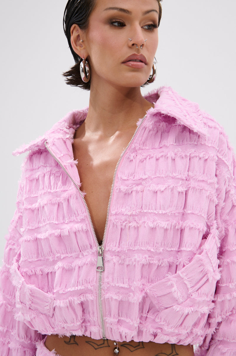 A woman with slicked-back hair and facial piercings wears the BUBBLEGUM BUBBLE HEM BOMBER, a textured pink jacket with distressed fringe, wide collar, and cropped silhouette. Silver hoops and chest tattoos complete her bold look.