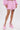A person wears textured pink BUBBLEGUM BUBBLE HEM SHORTS and a matching long-sleeve top, styled with ankle-strap platform heels and fringe trim accents, posing against a plain white background.