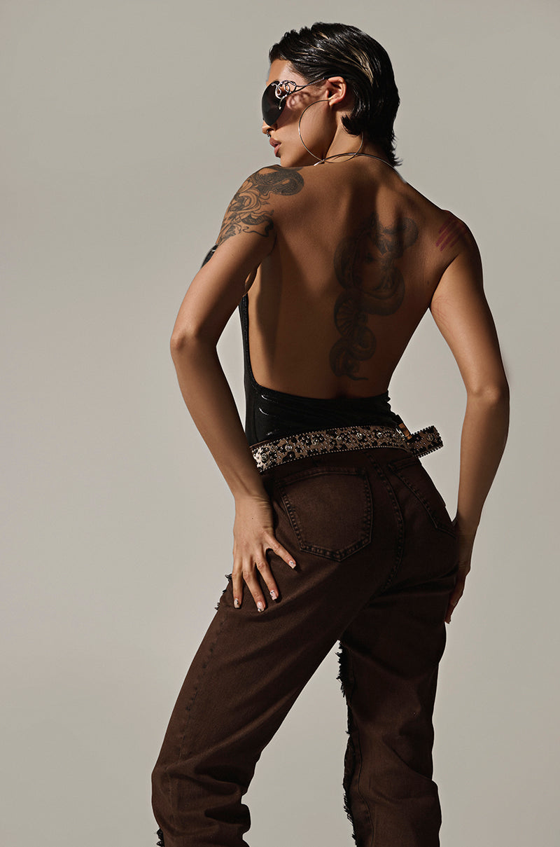 With short dark hair and tattoos, the individual poses confidently, their back to the camera in a curve-hugging fit wearing a sleeveless SLINKY FOIL BLACK BODYSUIT paired with brown pants and a studded belt. Adorned with large sunglasses and earrings, they stand against a plain gray background.