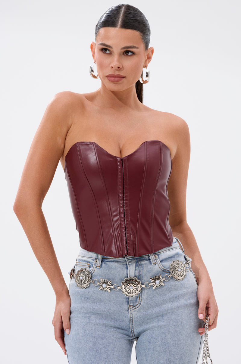 A woman models the BACK IT UP CORSET TOP IN BURGUNDY, a strapless faux leather piece, styled with light blue high-waisted jeans and a silver floral belt against a plain white background. 0