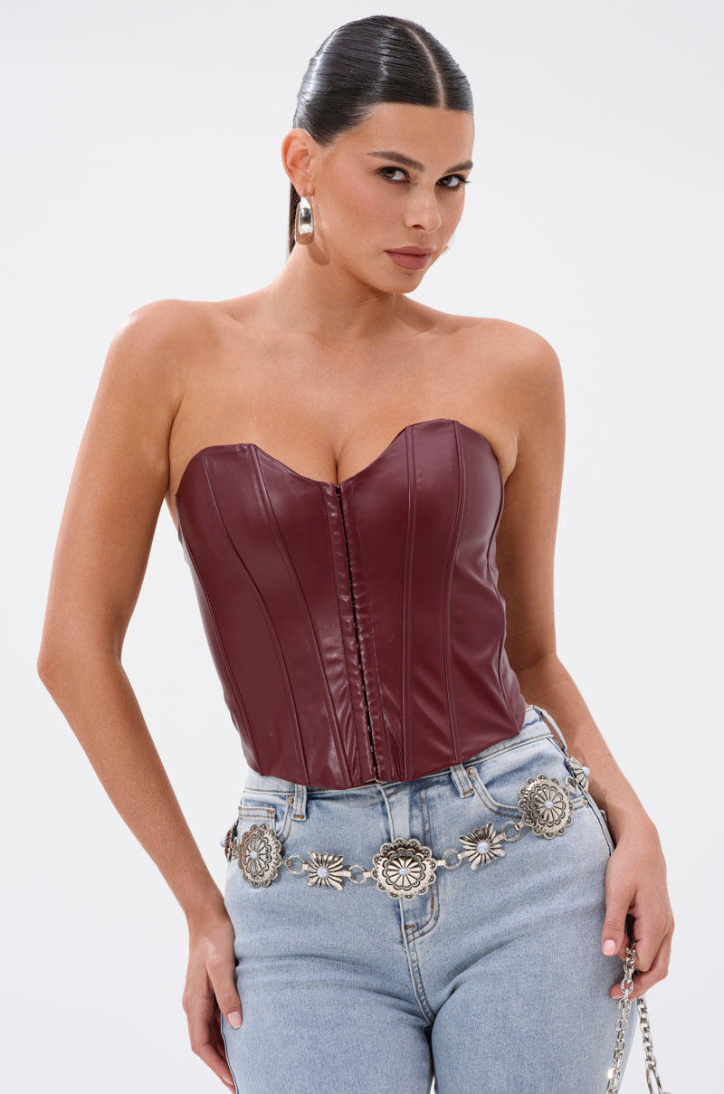 A woman with slicked-back dark hair poses confidently against a white background wearing the BACK IT UP CORSET TOP IN BURGUNDY, a strapless faux leather corset top, paired with silver hoop earrings and light blue jeans with a silver floral belt. 1