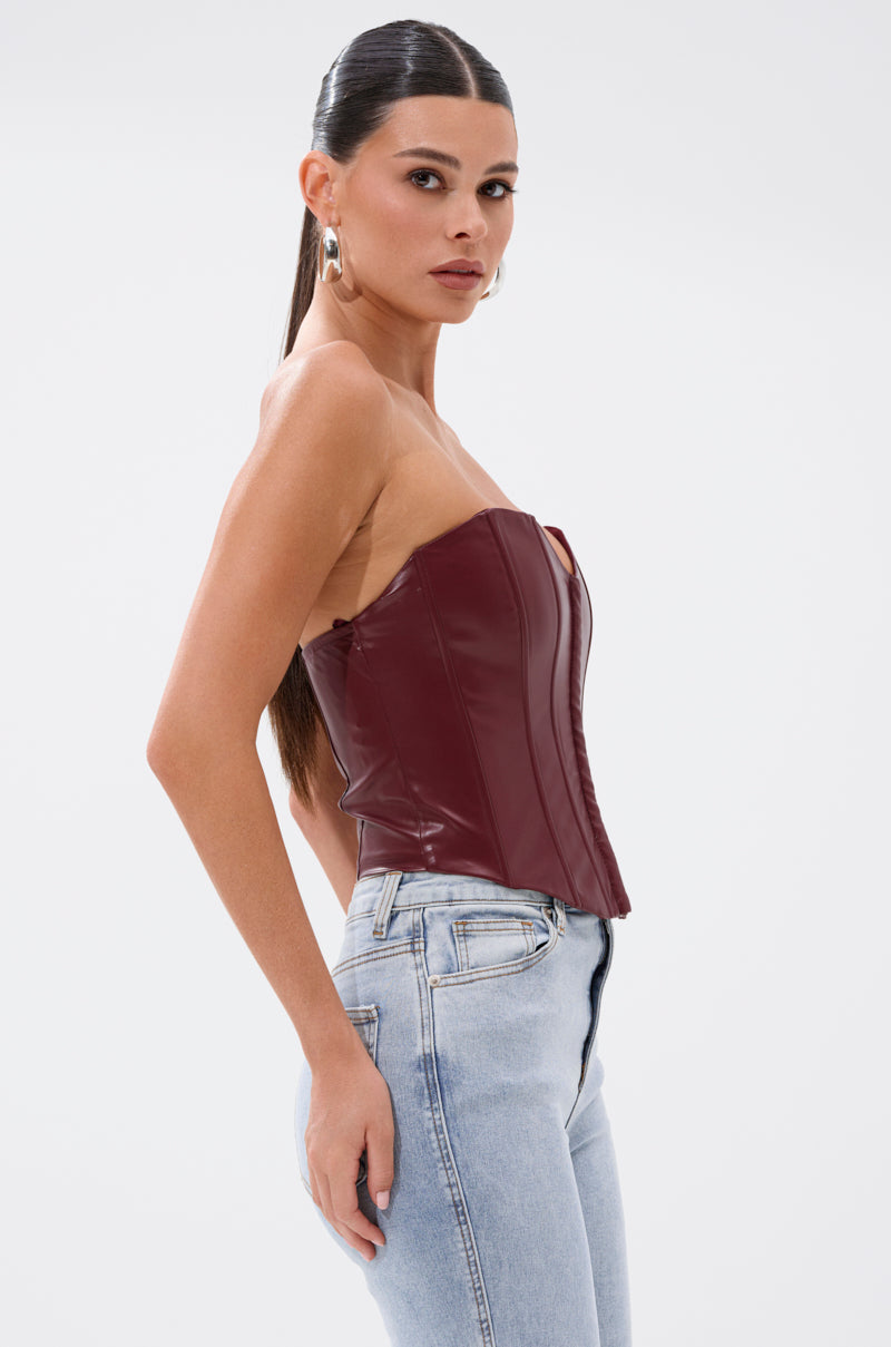 A woman with long dark hair in a ponytail models the BACK IT UP CORSET TOP IN BURGUNDY, paired with silver hoop earrings and high-waisted light-wash jeans, posing in profile against a plain white background. 2
