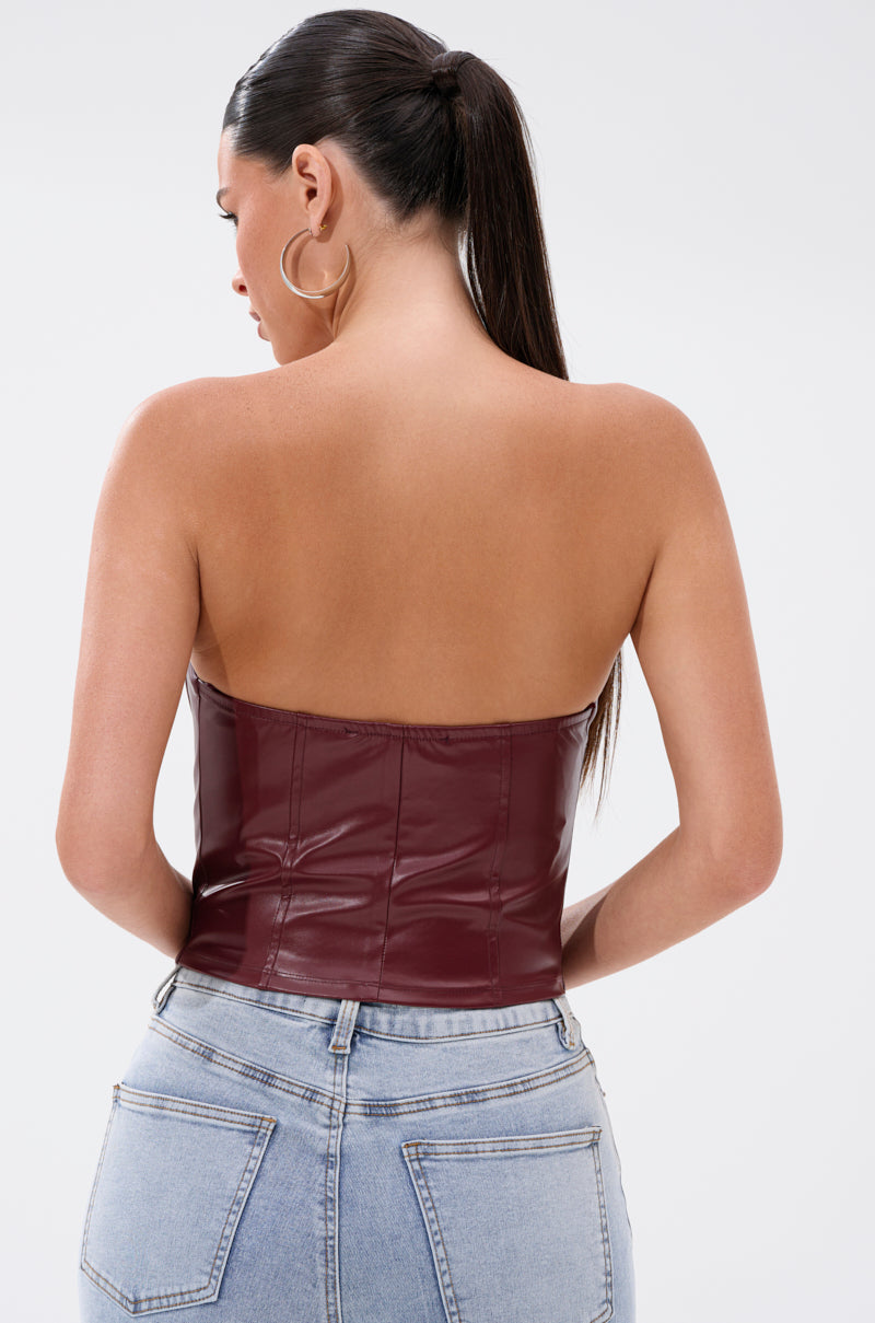 A woman with dark hair in a ponytail wears large hoop earrings and the BACK IT UP CORSET TOP IN BURGUNDY, a strapless faux leather piece, paired with light blue high-waisted jeans. She faces away, displaying the back of her outfit against a plain background. 3