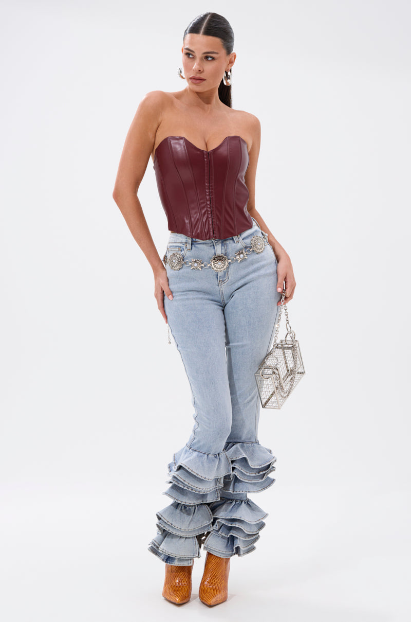 A woman wears the BACK IT UP CORSET TOP IN BURGUNDY with light-wash ruffled jeans, a silver belt, brown heeled boots, and a silver geometric handbag while posing against a white background. 5