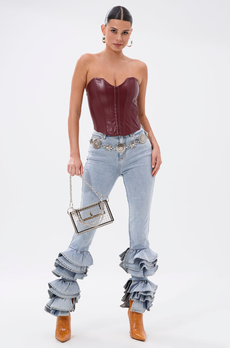A woman stands confidently in the BACK IT UP CORSET TOP IN BURGUNDY, paired with layered ruffle light blue jeans, brown ankle boots, a metallic belt, hoop earrings, and a small silver handbag. 6