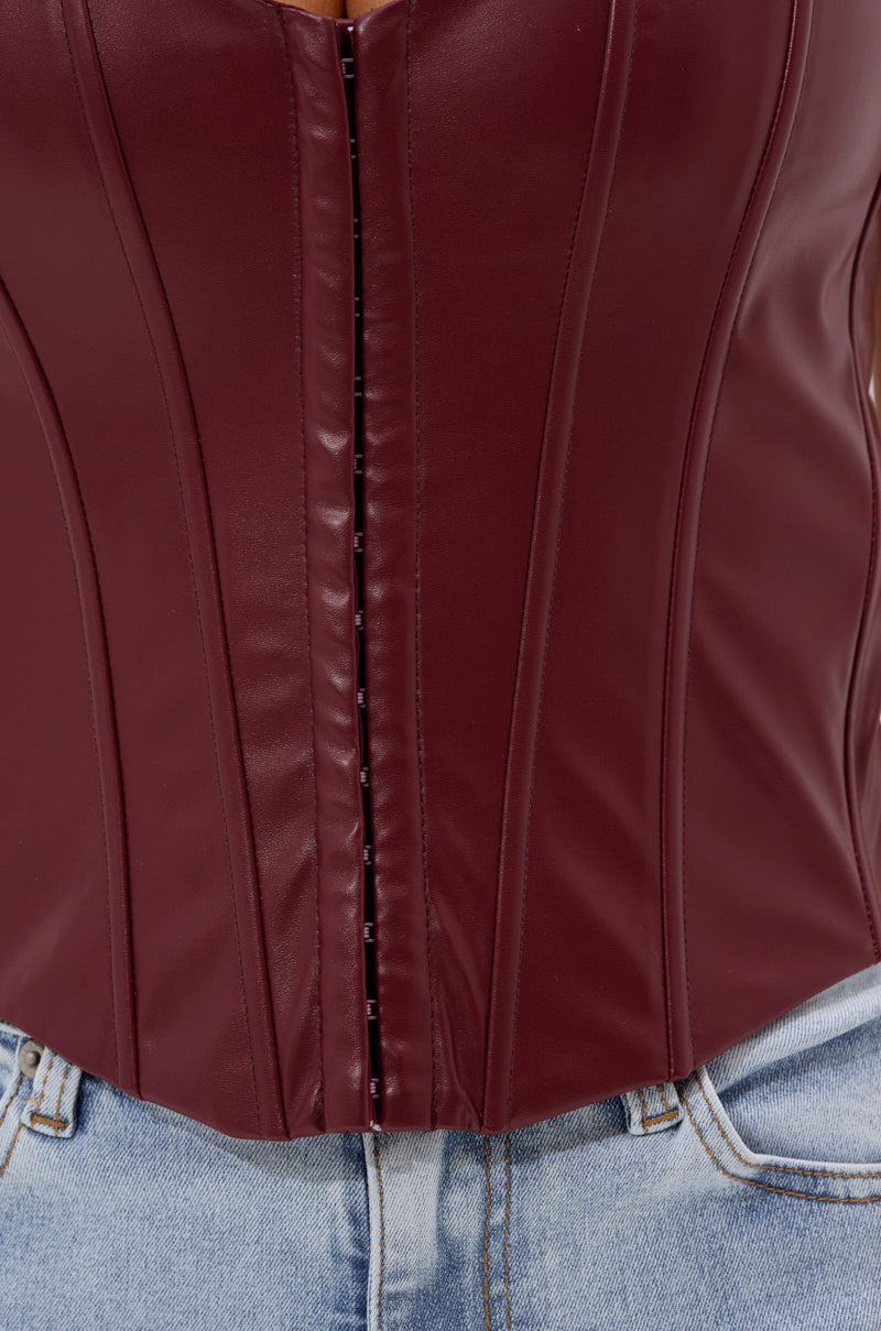 A close-up of a person wearing the BACK IT UP CORSET TOP IN BURGUNDY, crafted from faux leather with visible boning details, paired with light blue denim jeans. 7