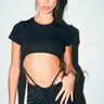 A woman with blue eyes and slicked-back hair wears the TOO SEXY FOR THIS BODYSUIT, paired with a high-waisted skirt with gold details, hoop earrings, and a gold clutch against a white brick wall.