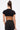 A woman with long brown hair in a high ponytail is seen from the back, wearing the TOO SEXY FOR THIS BODYSUIT—a black cut-out, open-back cropped top—paired with high-waisted black pants, gold accessories, and holding a small card.