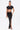A woman stands against a white background wearing the TOO SEXY FOR THIS BODYSUIT, paired with a high-slit, ruched black skirt with gold accents. She accessorizes with a gold clutch, black heels, and a high ponytail for a sleek look.