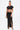 A woman poses against a white background wearing the "TOO SEXY FOR THIS BODYSUIT," a black cut-out design with side ring details, high slit, and short sleeves. She accessorizes with a gold clutch, high heels, and a sleek ponytail.
