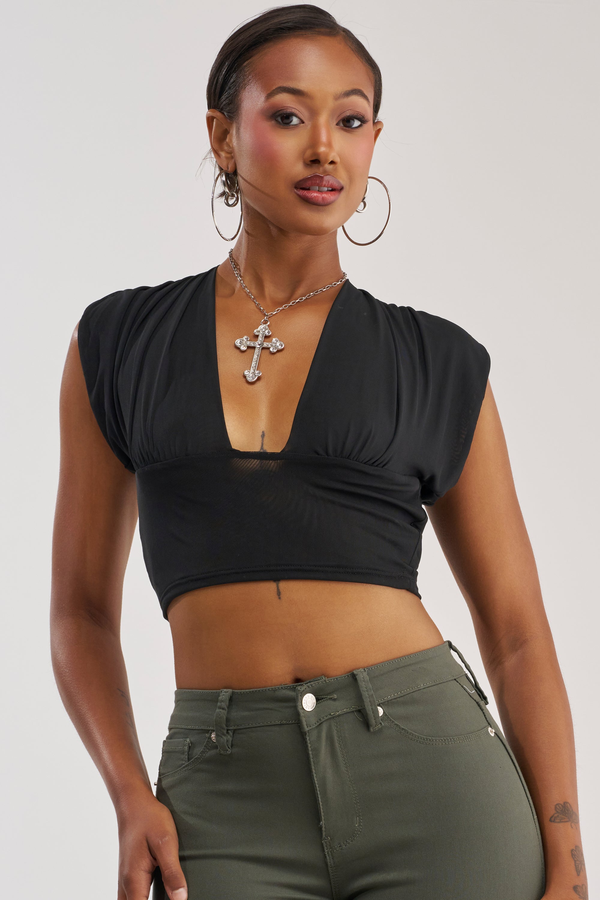 A woman stands confidently against a plain background, wearing the HOTTIE ALERT MESH BLOUSE—a sexy black deep V-neck crop top styled like a plunge blouse—paired with green pants, large hoop earrings, and a cross necklace as she looks at the camera.