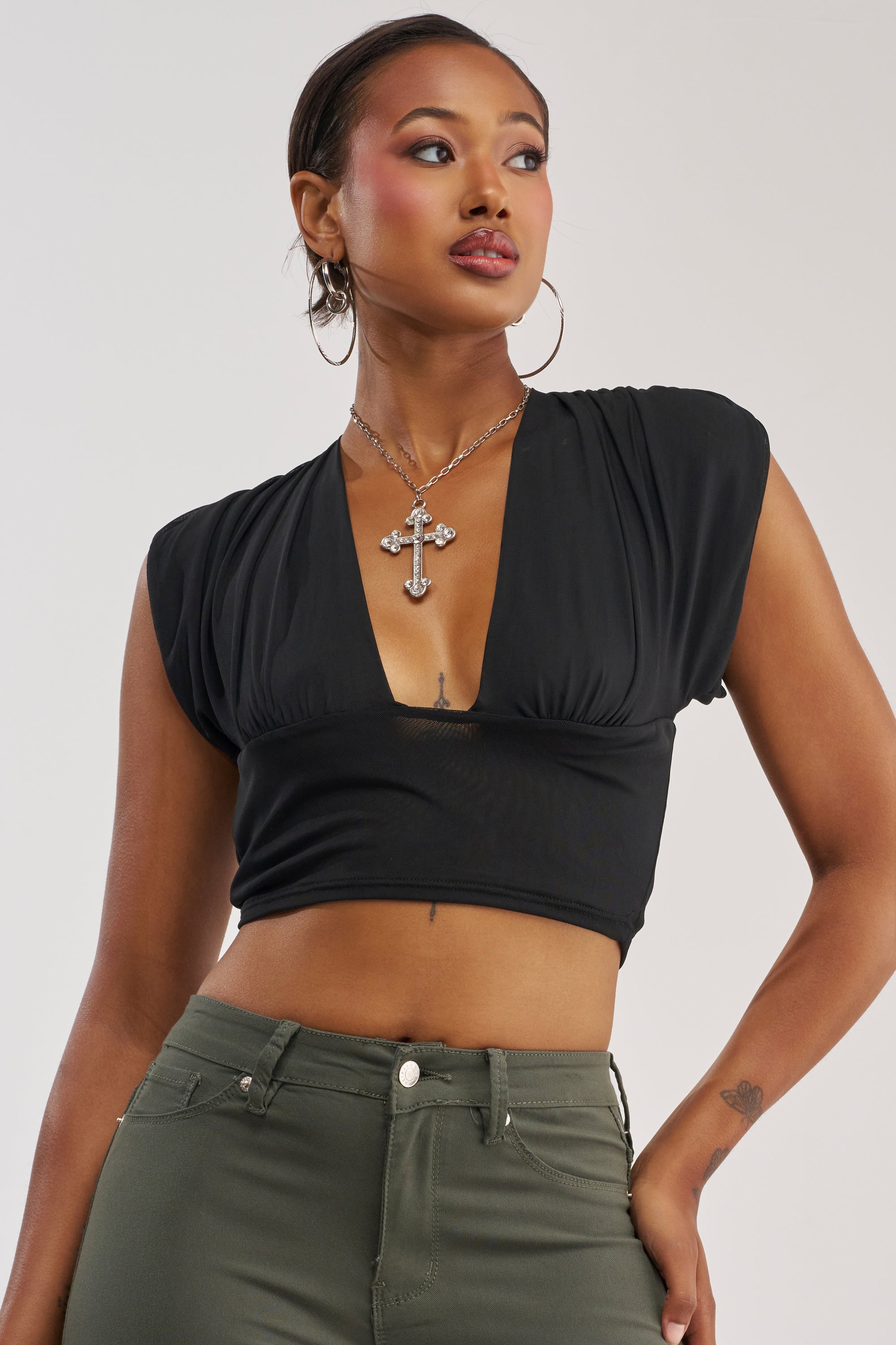 A woman stands against a light background with her right hand on her hip, wearing the HOTTIE ALERT MESH BLOUSE—a black, deep V-neck plunge blouse—paired with olive green pants, large hoop earrings, and a silver cross necklace.