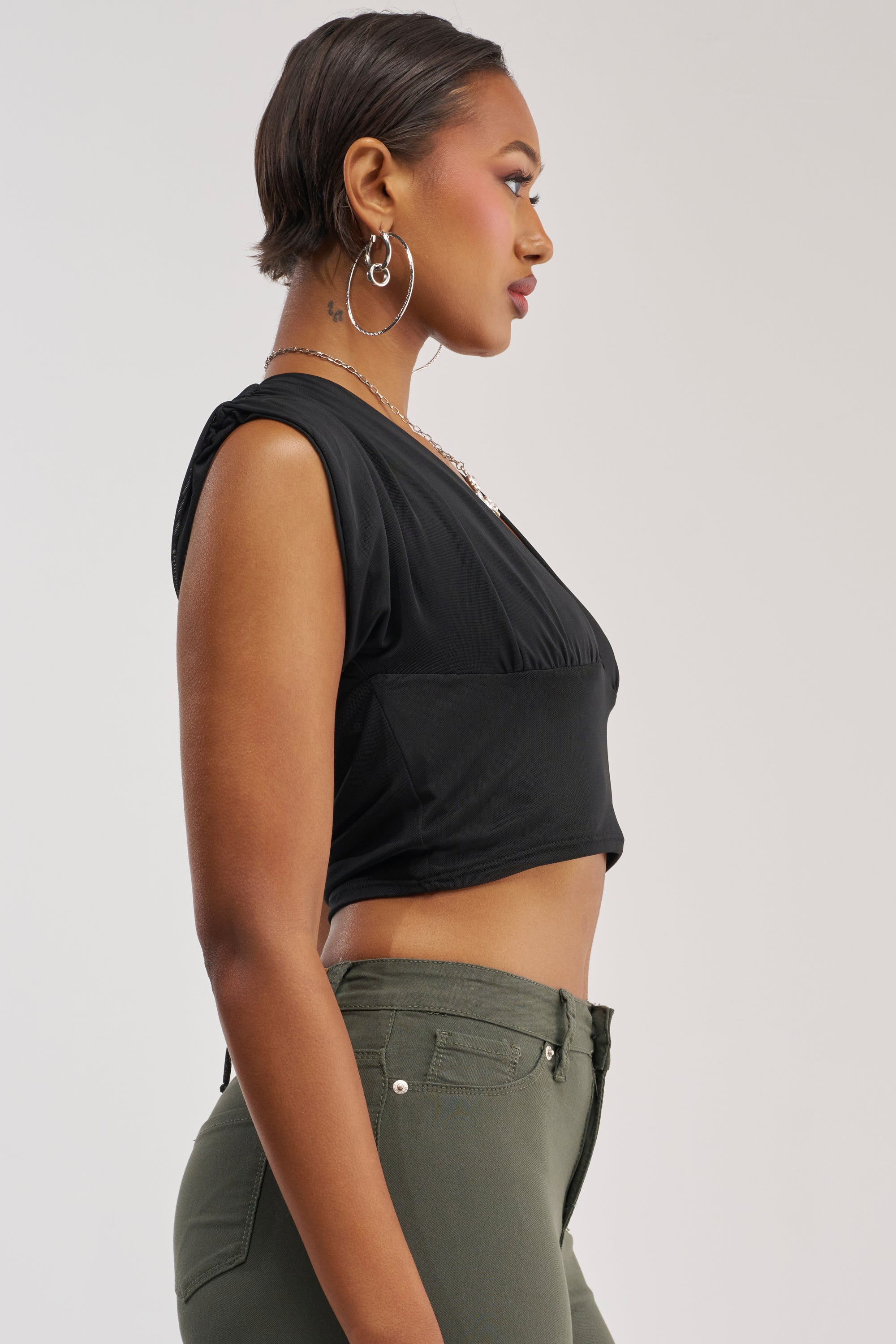 A woman stands in profile, wearing the HOTTIE ALERT MESH BLOUSE—a black sleeveless crop top—paired with olive green pants. She has short hair, hoop earrings, and a necklace, set against a light background.