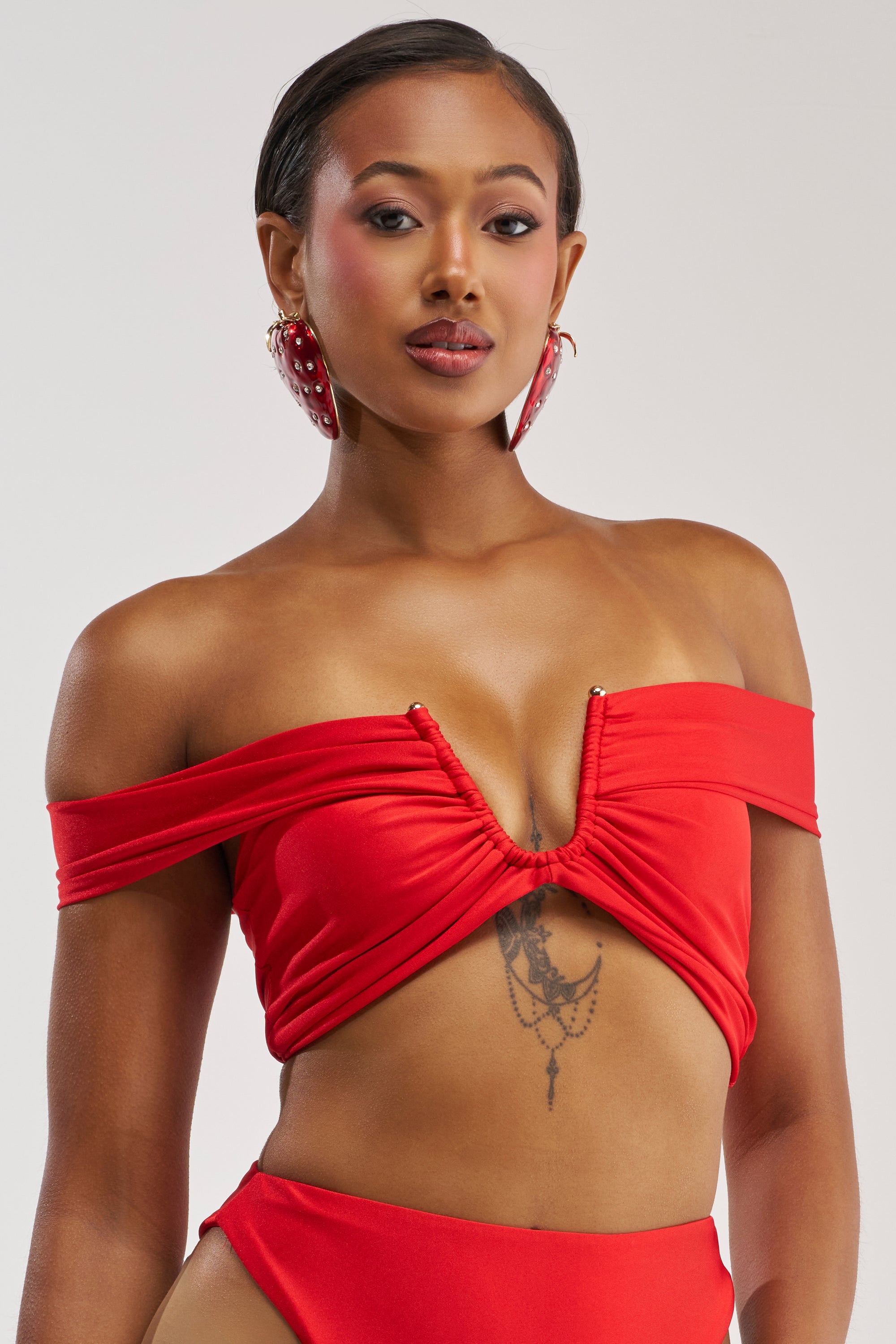 A woman poses confidently against a plain background wearing the HOTTIE ALERT MESH BLOUSE IN RED, styled off-the-shoulder with a ruched bodice and complemented by large red earrings, matching bottoms, and a chest tattoo.