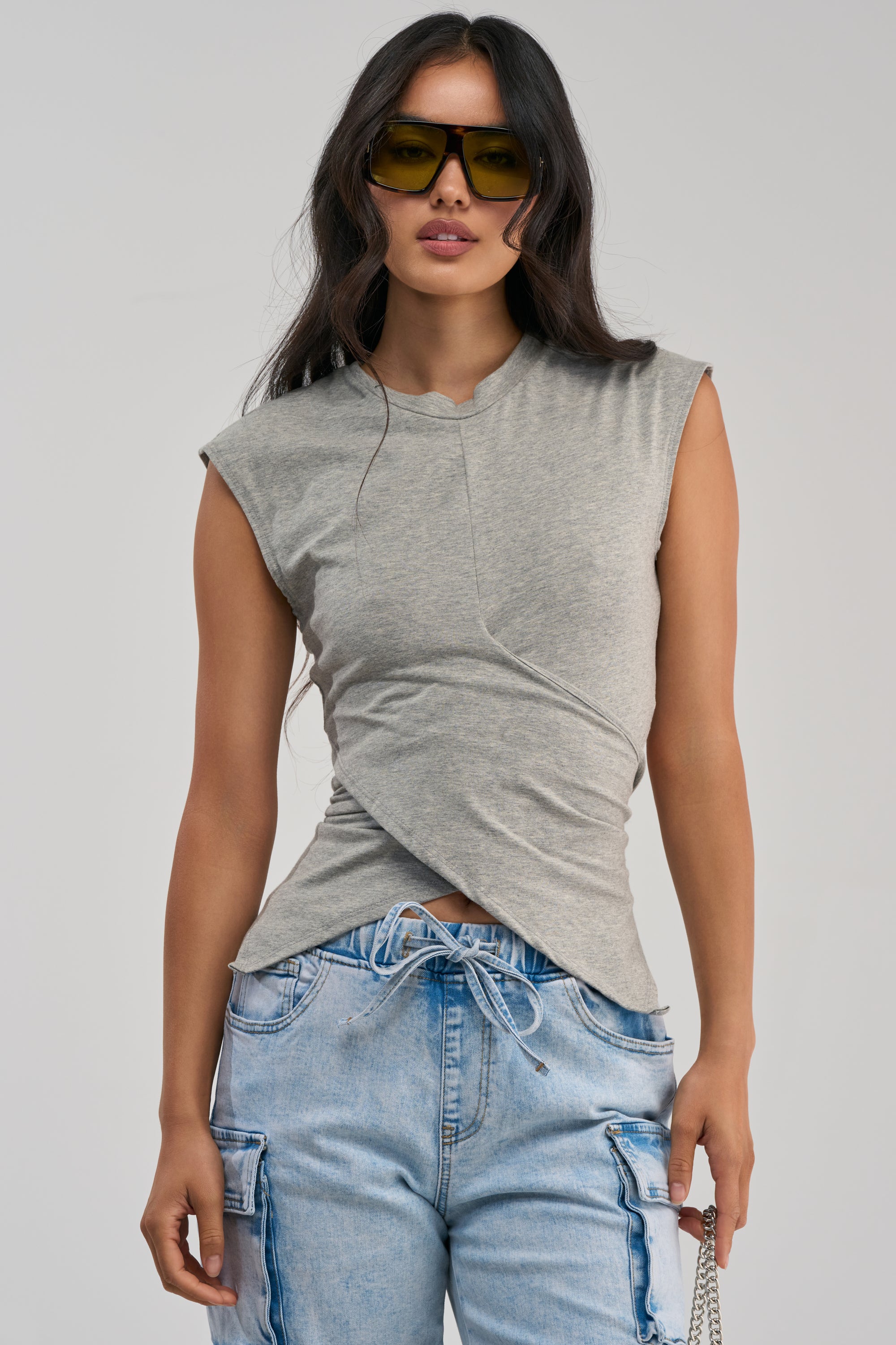A woman models the JUSTINE WRAP TEE—a fitted grey wrap-front top with cap sleeves—paired with light blue drawstring cargo jeans for an effortlessly stylish streetwear look. She completes her outfit with yellow sunglasses and a small chain purse, standing in front of a plain gray background.