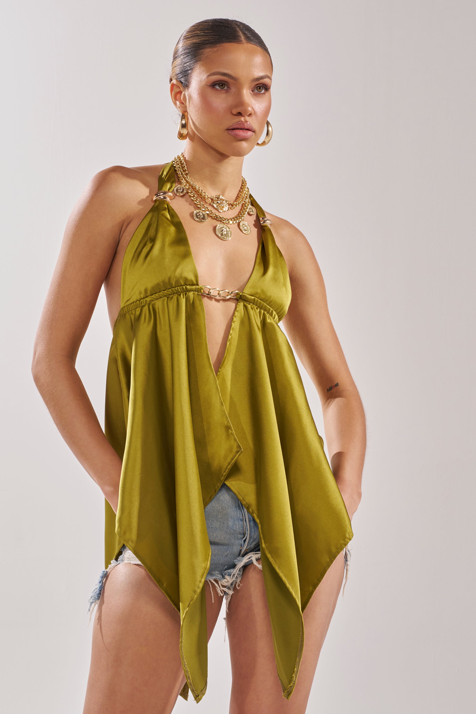 Wearing the LOTS OF LOVE TO GIVE HALTER NECK BLOUSE IN CHARTREUSE with distressed denim shorts and layered gold necklaces, she creates a chic going-out look against a plain backdrop. 0